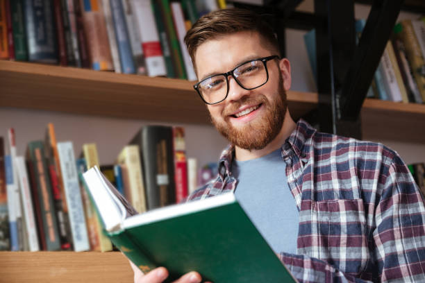 istock Smiling young male student in eyeglasses holding open book 1349197756