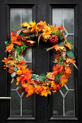A handmade, autumnal, halloween wreath hanging on a black door. The wreath is made up of leaves, flowers, acorns and small pumpkins and squashes.