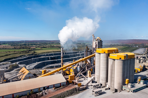 Aerial view of cement factory in industrial production area. Smoke is coming out of the chimney. It is a beautiful sunny autumn day and the leaves in the trees are multi colored. The factory is located close to agricultural fields.