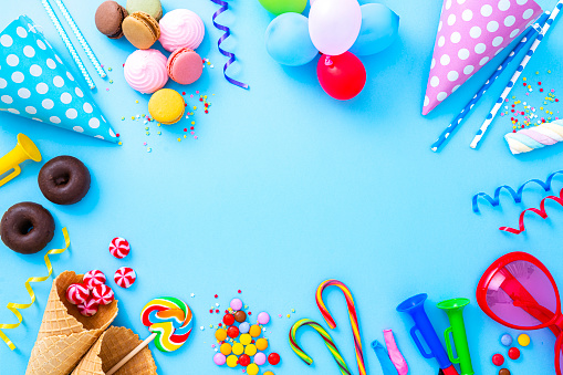 Party or carnival backgrounds. Overhead view of multi colored accessories like balloons, confetti, candies, party hat and drinking straws arranged all around a blue background making a frame and leaving useful copy space for text and/or logo. High resolution 42Mp studio digital capture taken with SONY A7rII and Zeiss Batis 40mm F2.0 CF lens