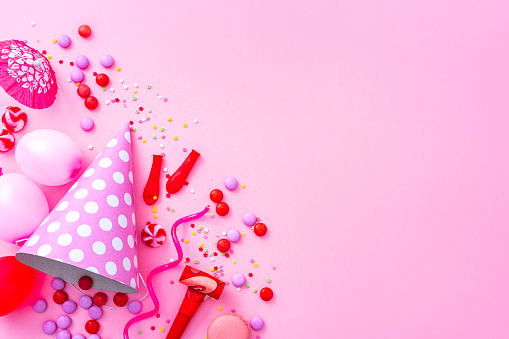 Party or carnival backgrounds. Overhead view of pink and red colored accessories like balloons, confetti, candies, party hat and streamers shot at the left of a blue background leaving useful copy space for text and/or logo. High resolution 42Mp studio digital capture taken with SONY A7rII and Zeiss Batis 40mm F2.0 CF lens