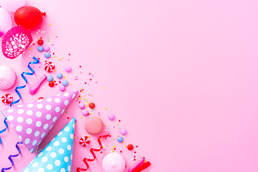 Party or carnival pink background. Overhead view of blue and pink colored accessories like balloons, confetti, candies, party hat and drinking straws shot at the top of a pink background making a frame and leaving useful copy space for text and/or logo. High resolution 42Mp studio digital capture taken with SONY A7rII and Zeiss Batis 40mm F2.0 CF lens