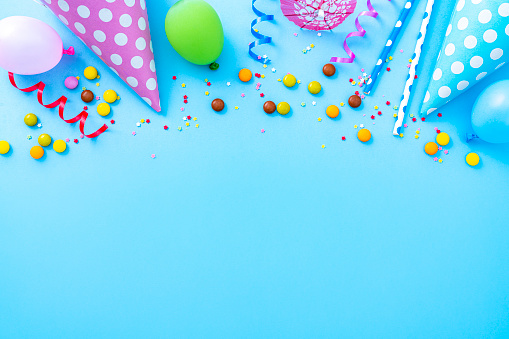 Party or carnival backgrounds. Overhead view of multi colored accessories like balloons, confetti, candies, party hat and drinking straws shot at the top of a blue background making a frame and leaving useful copy space for text and/or logo. High resolution 42Mp studio digital capture taken with SONY A7rII and Zeiss Batis 40mm F2.0 CF lens