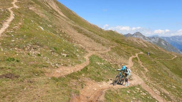 Woman mountain bikes along sunny mountain ridge She follows trail along mountain ridge, in the European Alps top view on people walking and cyclists riding stock pictures, royalty-free photos & images