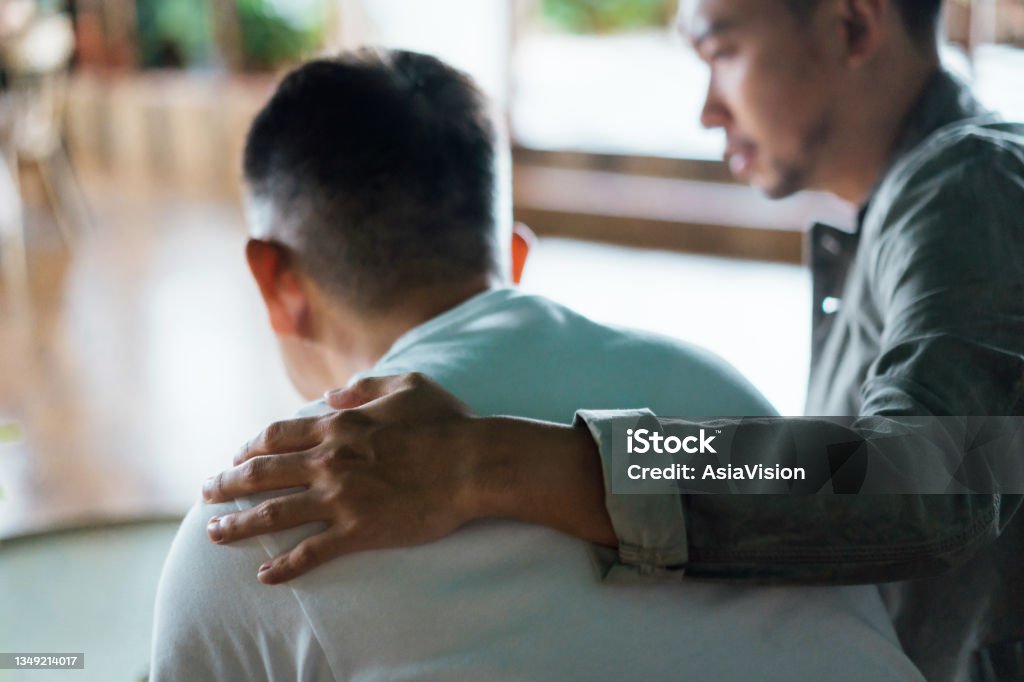 Rear view of son and elderly father sitting together at home. Son caring for his father, putting hand on his shoulder, comforting and consoling him. Family love, bonding, care and confidence Mental Health Stock Photo