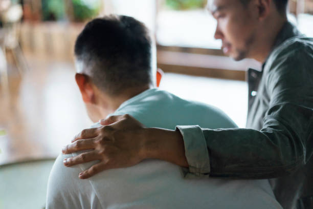 Rear view of son and elderly father sitting together at home. Son caring for his father, putting hand on his shoulder, comforting and consoling him. Family love, bonding, care and confidence stock photo