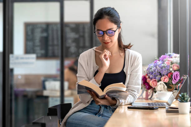 young asian woman holding a pencil reading a book with laptop smartphone on the desk. - woman with glasses reading a book imagens e fotografias de stock