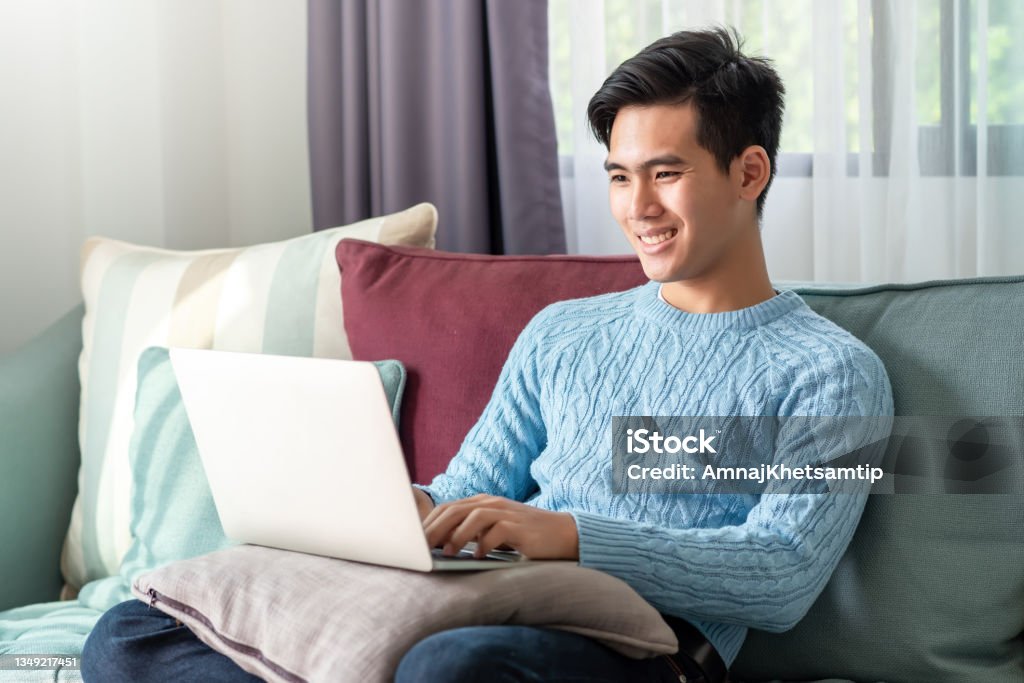 Happy of a leisurely day of a young Asian man watching his laptop, sitting on the sofa at home. Asia Stock Photo