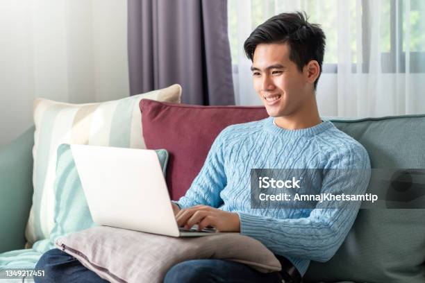 Happy Of A Leisurely Day Of A Young Asian Man Watching His Laptop Sitting On The Sofa At Home Stock Photo - Download Image Now