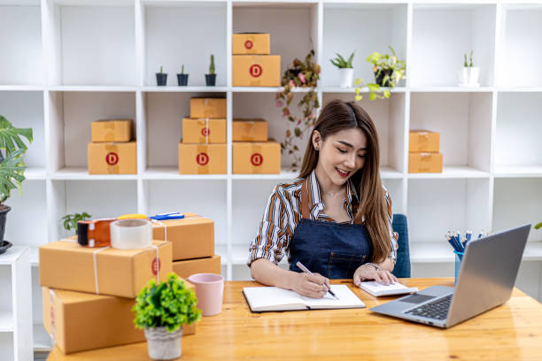 A beautiful Asian business owner opens an online store, she is checking orders from customers via laptop, sending goods through a courier company, concept of a woman opening an online business. una hermosa dueña de un negocio asiático abre una tienda en línea, está verificando los pedidos de los clientes a través de una computadora portátil, enviando productos a través de una empresa de mensajería, concepto de una mujer que abre un negoci