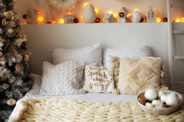 istock Cozy comfortable bed with Christmas pillows. White stylish scandinavian interior design room decorated with glowing garlands and fir tree for Xmas. Bedroom decor decorated new year or winter holiday. 1349221979