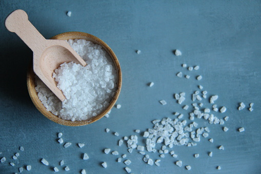 Sea salt in a wooden bowl isolated with a wooden spoon to measure on a light blue background, top view.