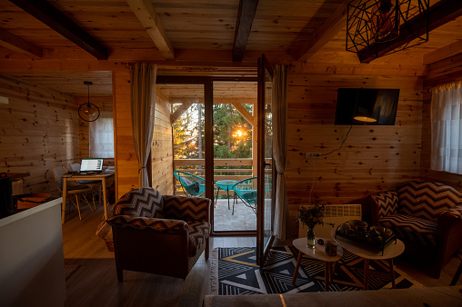 The interior design of a living room with large windows in a cozy rustic log cabin. Amazing view from the balcony of the sunset through the forest