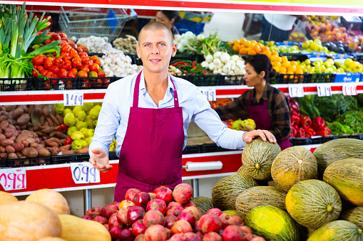Friendly smiling seller meeting customers in greengrocery store, offering fresh fruits and vegetables for sale