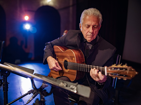 Mature male Guitarist on the stage with classic guitar. Grey hair. He is wearing all black. Stage interior of Theatre during performance at night.