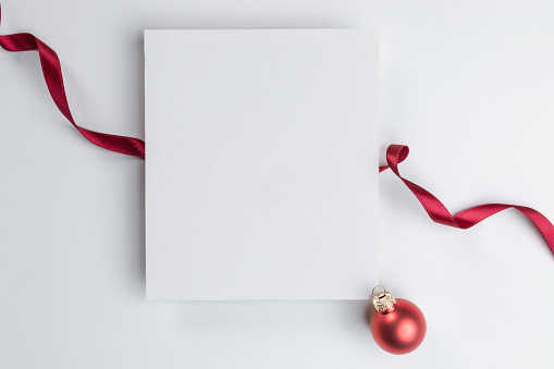 White blank card on white background wrapped with a red ribbon. At the right bottom corner of the blank card is one red Christmas ball.