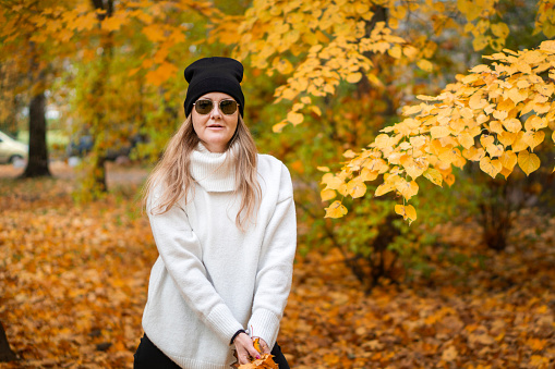 A photo of a pretty woman of mature age, smiling at the camera, wearing a hat and dark sunglasses, yellowed falling leaves, a walk and a beautiful unforgettable scent of autumn, city park.