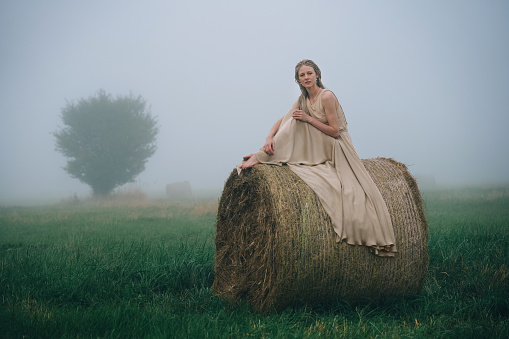 Young woman in beige dress sitting on haystacks in mist field at twilight time