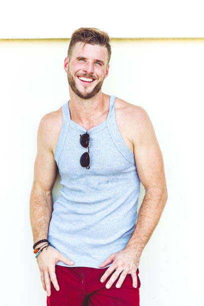 Portrait of a muscular man with a beard, seated indoors on a vintage sofa, wearing a white tank top. He stares directly at the camera with a serious, confident expression, arms crossed. The lighting is dramatic and gritty, adding an intense and raw visual atmosphere.