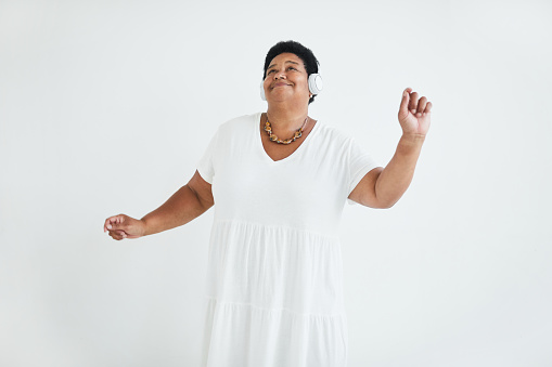 Happy senior woman in white dress dancing while listening to music in wireless headphones against the white background
