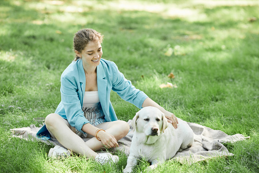 Full length portrait of smiling young woman playing with dog outdoors while enjoying walk together in park, copy space