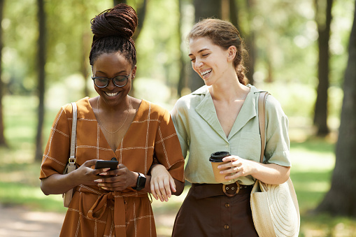 Waist up portrait of two smiling young women chatting outdoors while enjoying walk in park together