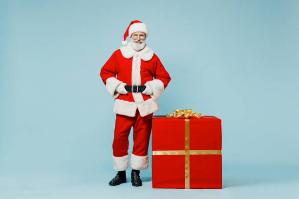 Full body old Santa Claus man 50s in Christmas hat red suit clothes stand akimbo near big gift box isolated on plain blue background studio. Happy New Year 2022 celebration merry ho x-mas concept. hombre de papá noel de cuerpo completo de 50 años con sombrero de navidad ropa de traje rojo de pie akimbo cerca de una gran caja de regalo aislada en un estudio de fondo azul liso. feliz año nuevo 2022 celebración feliz ho concepto de navidad