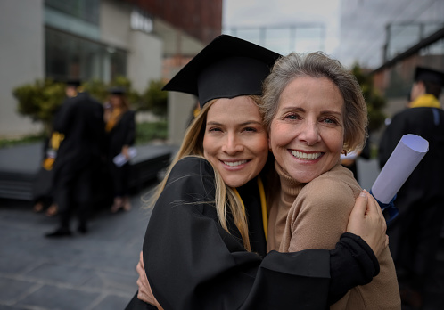 Portrait of a happy graduate student hugging her mother on graduation day and looking at the camera smiling - education concepts