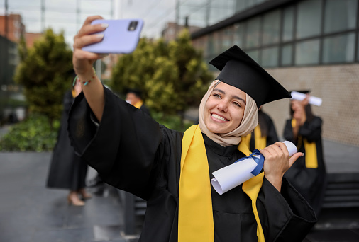 Happy Muslim female graduate student taking a selfie with her diploma on her graduation day using a cell phone - education concepts
