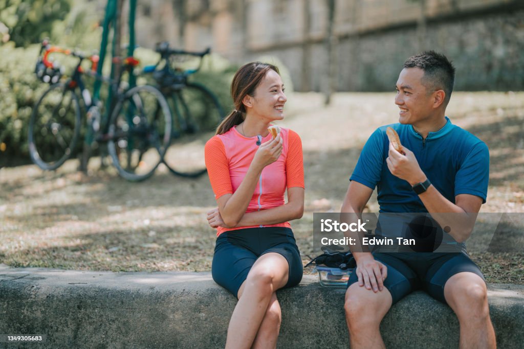 Asian chinese couple cyclist taking a break enjoying sandwiches at river front after morning road trip cycling in public park Eating Stock Photo