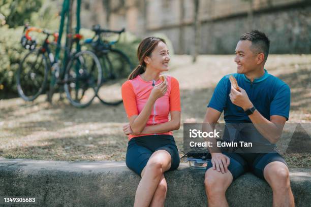 Asian Chinese Couple Cyclist Taking A Break Enjoying Sandwiches At River Front After Morning Road Trip Cycling In Public Park Stock Photo - Download Image Now