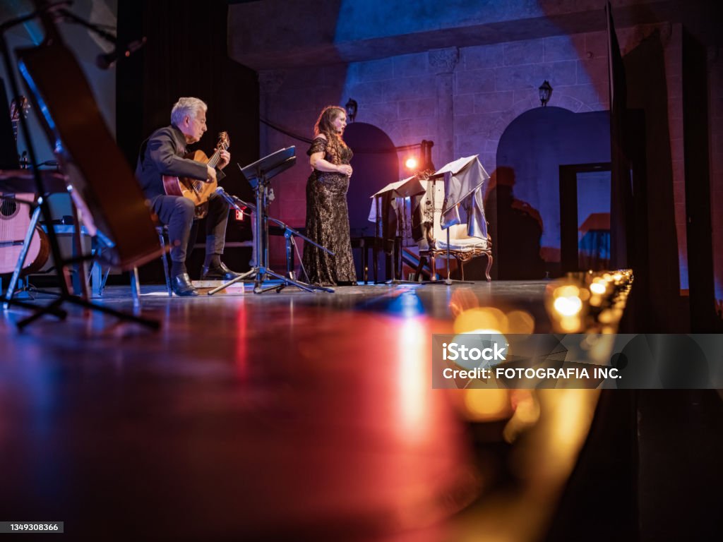 Mature Performer and Guitarist on the stage Mature woman Performer and male Guitarist on the stage. She is dressed in period costume, he is wearing all black. Stage interior of Theatre during performance at night. Performance Group Stock Photo