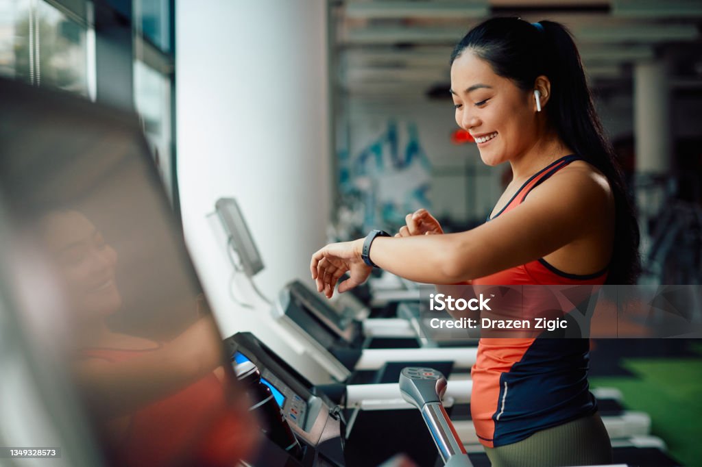 Happy Asia athletic woman using fitness tracker while running on treadmill in a gym. Happy Asian sportswoman using smart watch while jogging on treadmill during sports training in a gym. Fitness Tracker Stock Photo