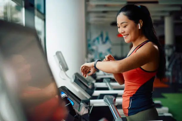 Happy Asian sportswoman using smart watch while jogging on treadmill during sports training in a gym.