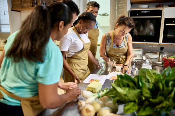 Adult Students Reading Recipe in Cooking Class Side view of diverse group of men and women wearing aprons and standing at kitchen workstation as they participate in team building event. cooking class photos stock pictures, royalty-free photos & images
