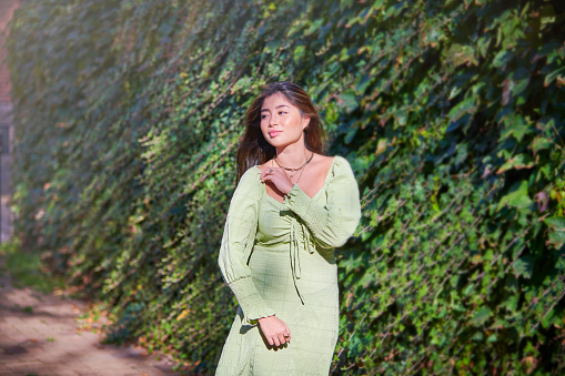 Model outdoors in city / street. Young asian ethnicity woman  with long hair smiling to the camera. Model is posing in front of big green plant wall