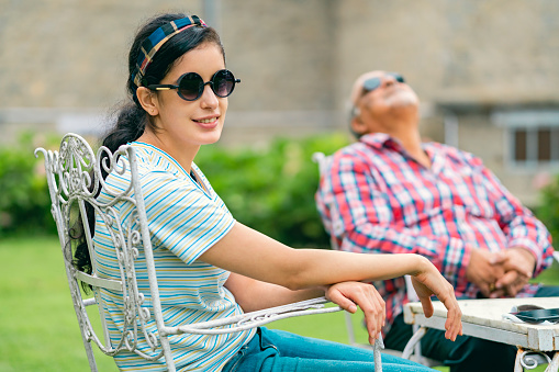 In this outdoor image, an Asian/Indian young woman looks away with happiness sitting in the daytime on the lawn chair with her father relaxing in the background during their vacation. They wear dark sunglasses.