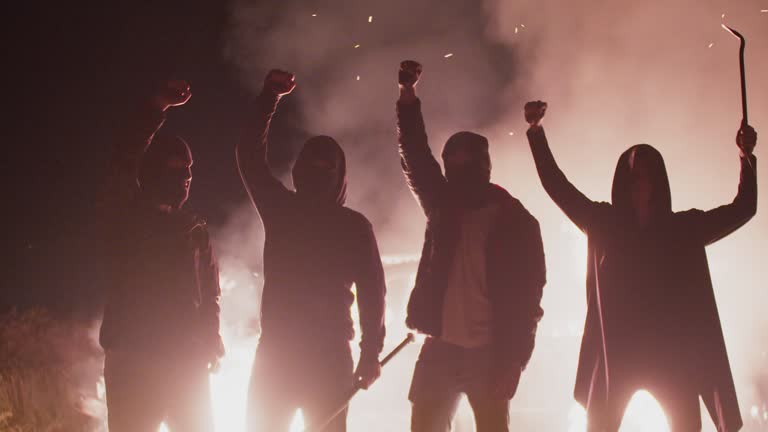 Four silhouettes of rebels in black masks with various edged weapons and with raised fists stand background of burning car, front view. Anarchists set fire to car in protest. Concept of protest