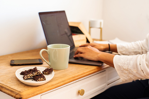 Close-up of a young woman having a coffee and snack while working remotely from home with a laptop