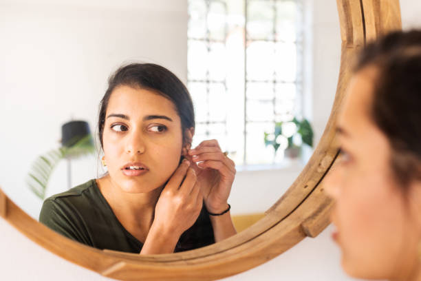 young woman putting on earrings in a mirror at home - brinco imagens e fotografias de stock