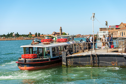 Murano, Italy - June 2, 2021: Ferry boats station of Murano island called Punta Faro with a group of people, Venice lagoon, UNESCO world heritage site. On background the San Michele island. Veneto, Italy, Europe. A group of tourists and locals just got off the vaporetto on a sunny spring day. The island of murano is famous for its industries that produce artistic glass.