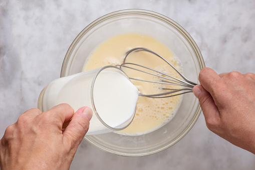 Top view of woman hand holding glass of milk putting into mixed raw eggs in glass bowl on marble surface