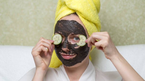 Girl in purifying black mask on face covering eyes by slices cucumber one of which is bitten. Charcoal face mask or black clay mud. Natural cosmetic concept. Skin care, acne treatment, cleansing skin. Girl in purifying black mask on face covering eyes by slices cucumber one of which is bitten. Charcoal face mask or black clay mud. Natural cosmetic concept. Skin care, acne treatment, cleansing skin. women facial mask mud cucumber stock pictures, royalty-free photos & images