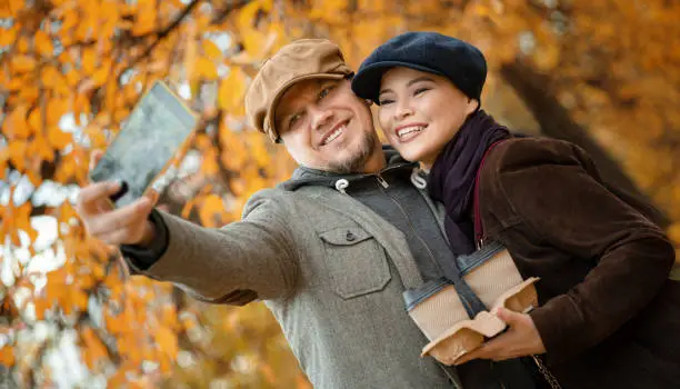 Smiling Couple With Coffee-to-go Makes Selfie in the Autumn City Park. Asian Woman With her Boyfriend Goes for a Walk in the Fall Forest in the Suburb. Close-up.
