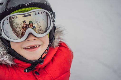 Mom and Dad are reflected in the boy ski goggles. Mom and Dad teach a boy to ski or snowboard.