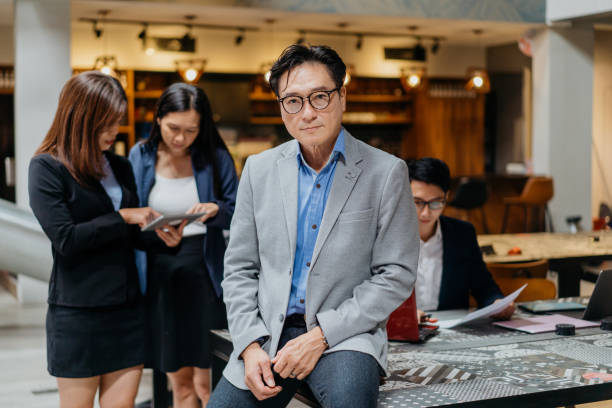 Confidence business leader and his team in office Portrait of a confidence Asian business leader arms leaning on table and looking at camera chairperson stock pictures, royalty-free photos & images