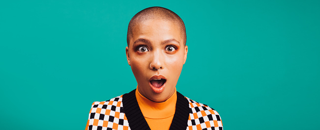 Woman looking shocked in a studio. Fashionable young woman looking at the camera with her eyes and mouth wide open. Hipster woman with a septum ring standing against a turquoise background.