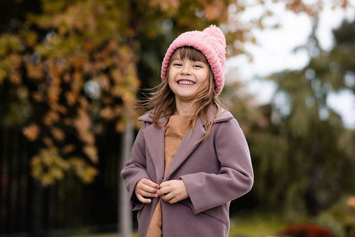 Happy cute child girl 4-5 year old wear knit wool hat and jacket over nature backggound outdoors. Autumn season. Looking at camera. Happiness. Laughing little kid having fun in park. Childhood.