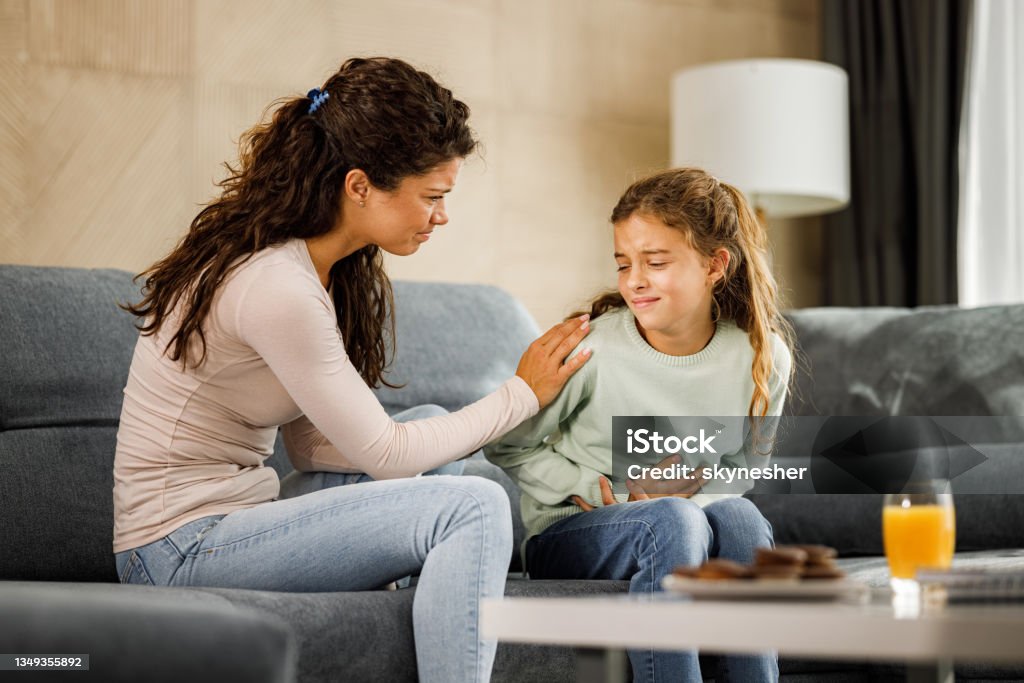 Mommy, my stomach hurts really bad! Worried single mother consoling her little girl who is having stomachache at home. Stomachache Stock Photo