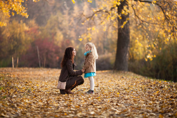 Mom sitting down and talking to her little daughter at eye level, holding her hands in the autumn park. stock photo